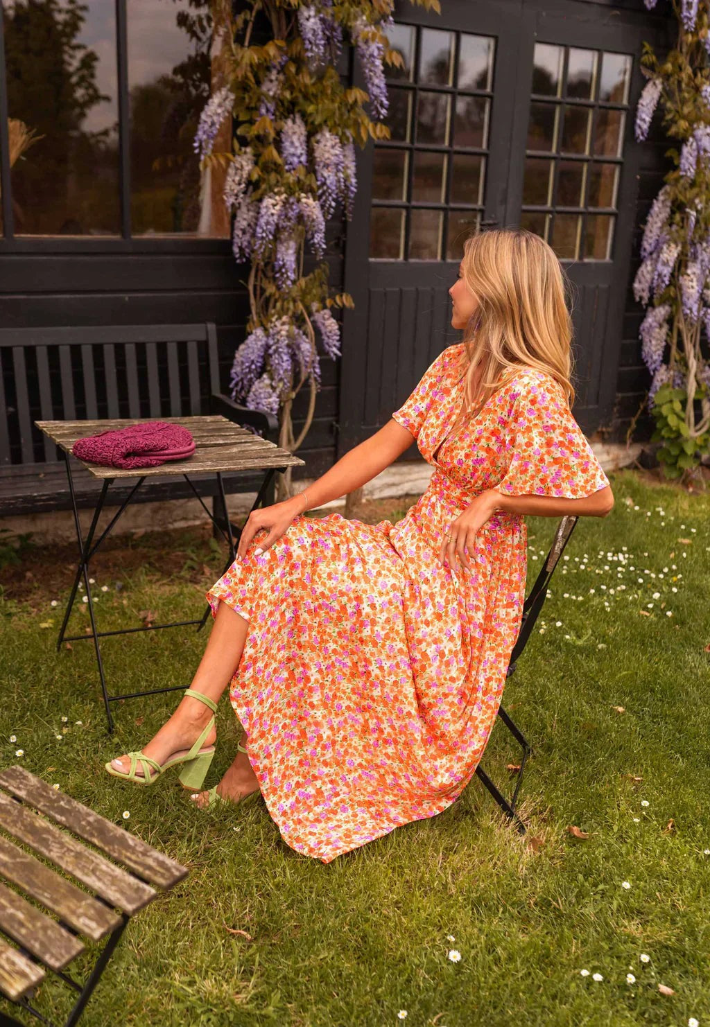 Orange Honoria women's floral wrap summer maxi dress, seated side view in a garden